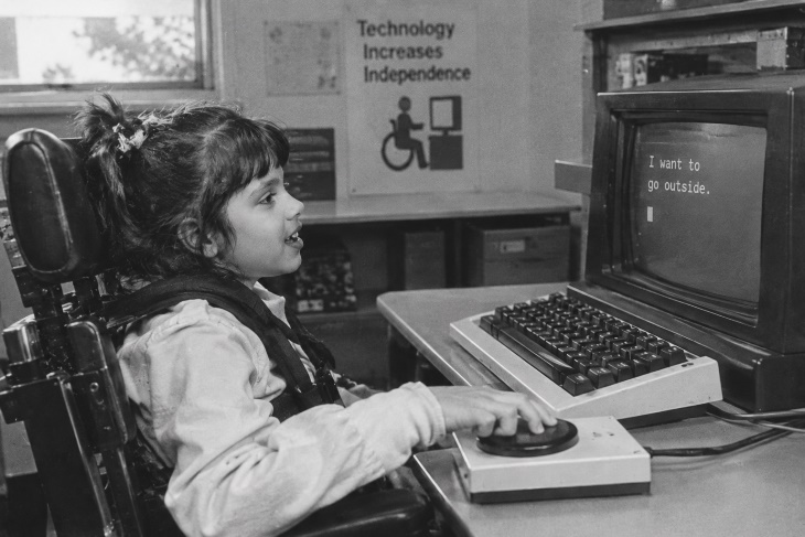 A young girl uses an adaptive joystick to work at a computer.